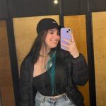 Woman with dark hair and a blue lock, wearing a black cap, black shirt and coat and jeans, smiling to a mirror and taking a picture with a purple phone.
