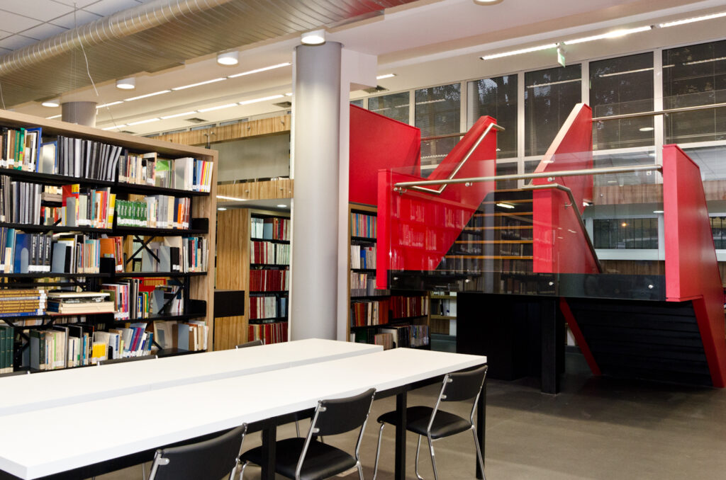 Image of a libriry, with a white table with black chair in the front, bookshelves in the back and a red and black stairs on the right side.