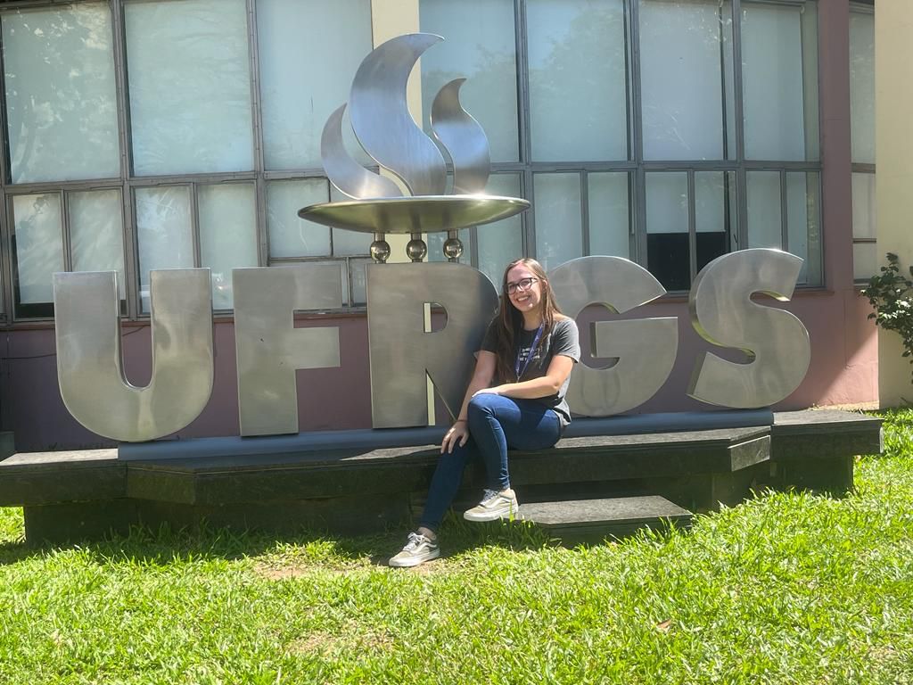 A blonde girl smiling towards the camera and sitting in front of the UFRGS monument.